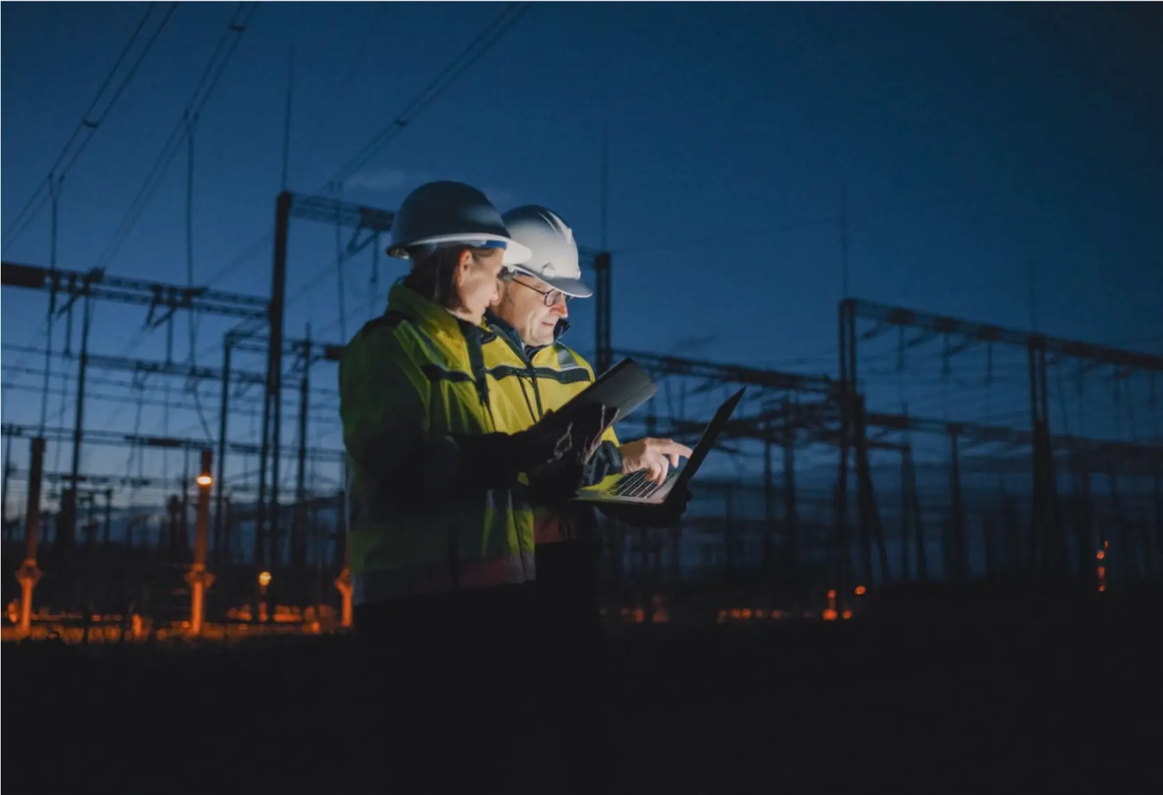 Two engineers in safety gear working on laptops at an outdoor power station at night