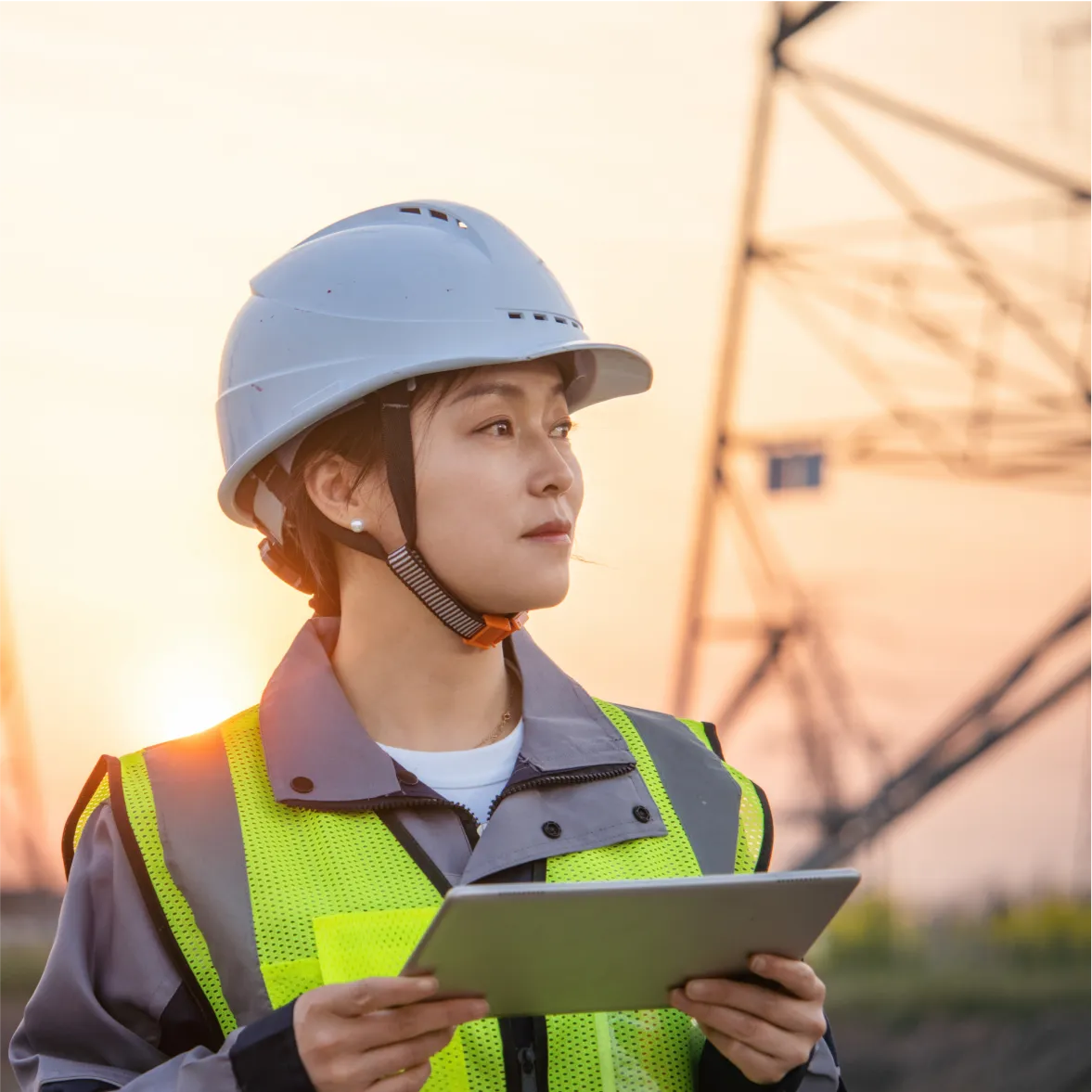 Female engineer with safety gear inspecting power lines at sunset using digital tablet